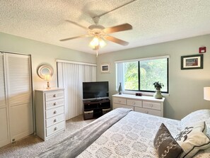 A bedroom with a bed, dresser, television, ceiling fan, window with view, and a clock. - BT 3821 Pond View Condo-Paradise Vacation Rental in Ocean Village (Fort Pierce)