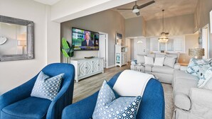 A spacious living room with a large flat-screen TV, a ceiling fan, and a dining area with a white cabinet and a plant. - CP 133 Ocean Front Condo - Vacation Rental in Ocean Village (Fort Pierce)