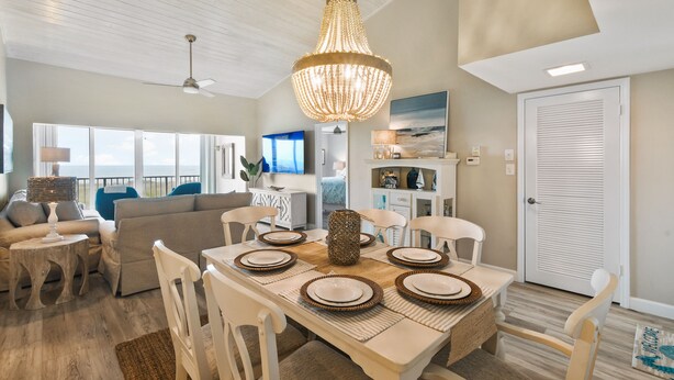 A dining area with a wooden table set for four, surrounded by white chairs. A chandelier hangs above the table, and a ceiling fan is visible in the background. - CP 133 Ocean Front Condo - Vacation Rental in Ocean Village (Fort Pierce)