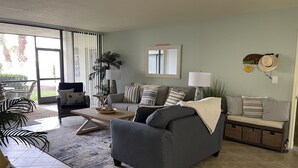 Ein Wohnzimmer mit grauem Sofa, einem Couchtisch und einem Streifen Teppich. Ein Fenster mit Vorhängen und eine Pflanze in der Ecke.