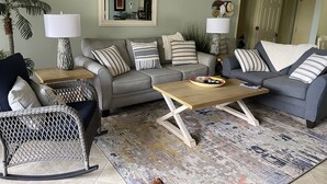 Ein Wohnzimmer mit grauem Sofa, einem Korbstuhl, einem Couchtisch und einem gemusterten Teppich.