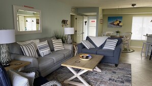 Ein Wohnzimmer mit grauem Sofa, einem Couchtisch und einem Korbstuhl. Es gibt einen Essbereich mit Tisch und Stühlen.