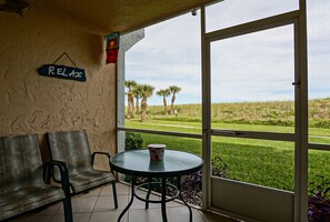 A room with a view of a grassy area and palm trees, a table with a cup, two chairs, and a 'RELAX' sign on the wall. - CP 114 Dune View Condo - Welcome to Paradise (Fort Pierce)