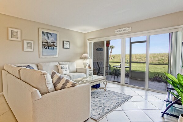 A living room with a beige sofa, a glass coffee table, and a view of the outdoors through large sliding glass doors. - CP 114 Dune View Condo - Welcome to Paradise (Fort Pierce)