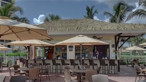 Un café en plein air avec des tables et des chaises placées sous de grands parasols, un coin bar doté d’un toit de chaume et, au fond, des palmiers.
