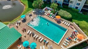 An aerial view of a hotel with a rectangular swimming pool surrounded by lounge chairs and umbrellas, a small pond, and a building with multiple windows. - CP 822 Golf Course View Condo - Paradise Vacation Rental in Ocean Village (Fort Pierce)