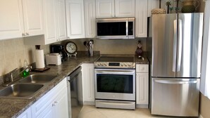 A modern kitchen with white cabinets, stainless steel appliances including a microwave, oven, and refrigerator, a granite countertop, and a small clock on the wall. - CT 2345- Ocean View Condo  -  in Ocean Village (Fort Pierce)