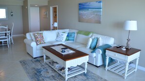 A living room with a white sofa, a wooden coffee table, and a side table with a lamp. There is a painting on the wall and a door leading to another room. - CT 2345- Ocean View Condo  -  in Ocean Village (Fort Pierce)