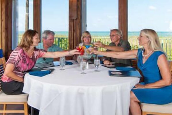 Een groep mensen die proosten aan een ronde tafel met een witte tafelkleed, met uitzicht op een strand.