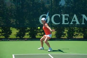 Een tennisbaan met een speler in actie, een hek en het woord "OCEAN" gedeeltelijk zichtbaar.