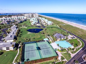 Vista aérea de um resort com vários edifícios, quadra de tênis e praia.
