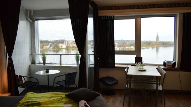 Habitación de hotel con cama, una mesa de comedor, una silla, un taburete pequeño y un ventanal con vistas a un cuerpo de agua y edificios.