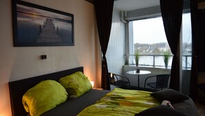 Habitación de hotel con cama, vistas a la ciudad a través de un ventanal grande, una mesita en el balcón y una silla.
