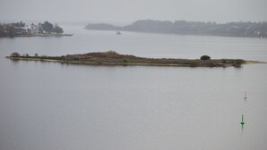 Un lago tranquilo con una pequeña isla en el centro, rodeado de edificios y una vista distante de un horizonte urbano.