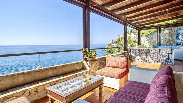 A coastal terrace with a stone wall, a wooden table, a cushioned sofa, and a bar area with stools. - SELENE - Eight Bedroom Villa, Sleeps 16 (Ravello)