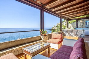 A coastal terrace with a stone wall, a wooden table, a cushioned sofa, and a bar area with stools. - SELENE - Eight Bedroom Villa, Sleeps 16 (Ravello)