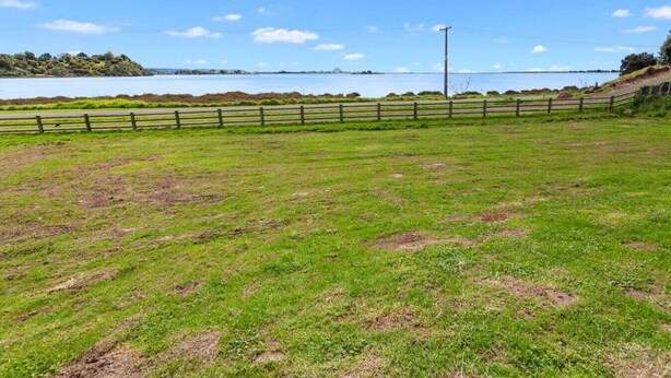 A grassy field with a wooden fence and a body of water in the background. - Kotare Koki · (Kingfisher Corner) (Opotiki)