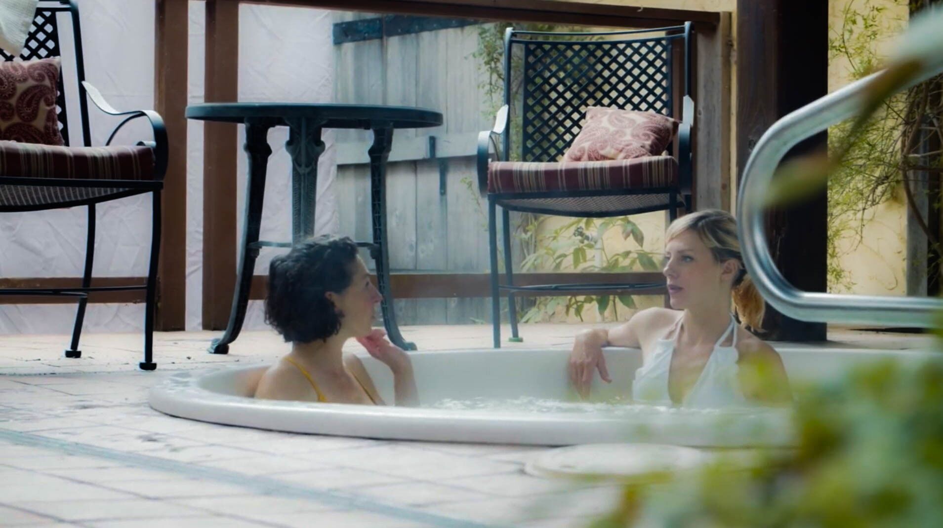 Un jacuzzi con dos personas, rodeado de mobiliario exterior y una valla de madera.