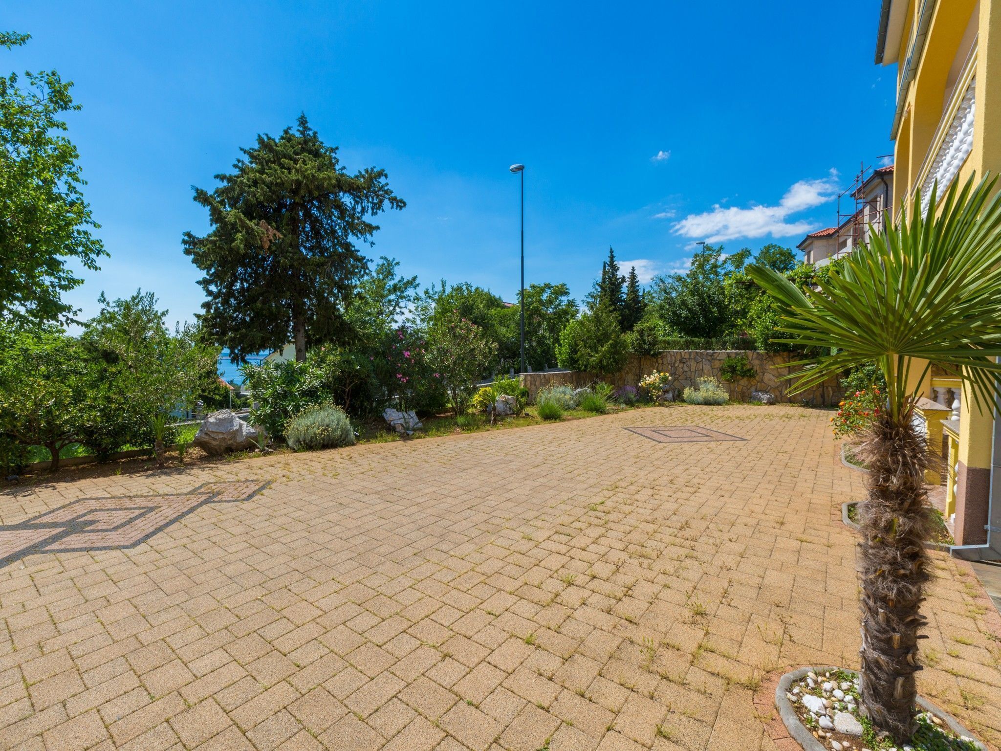 A paved courtyard with a palm tree, surrounded by greenery and a stone wall.