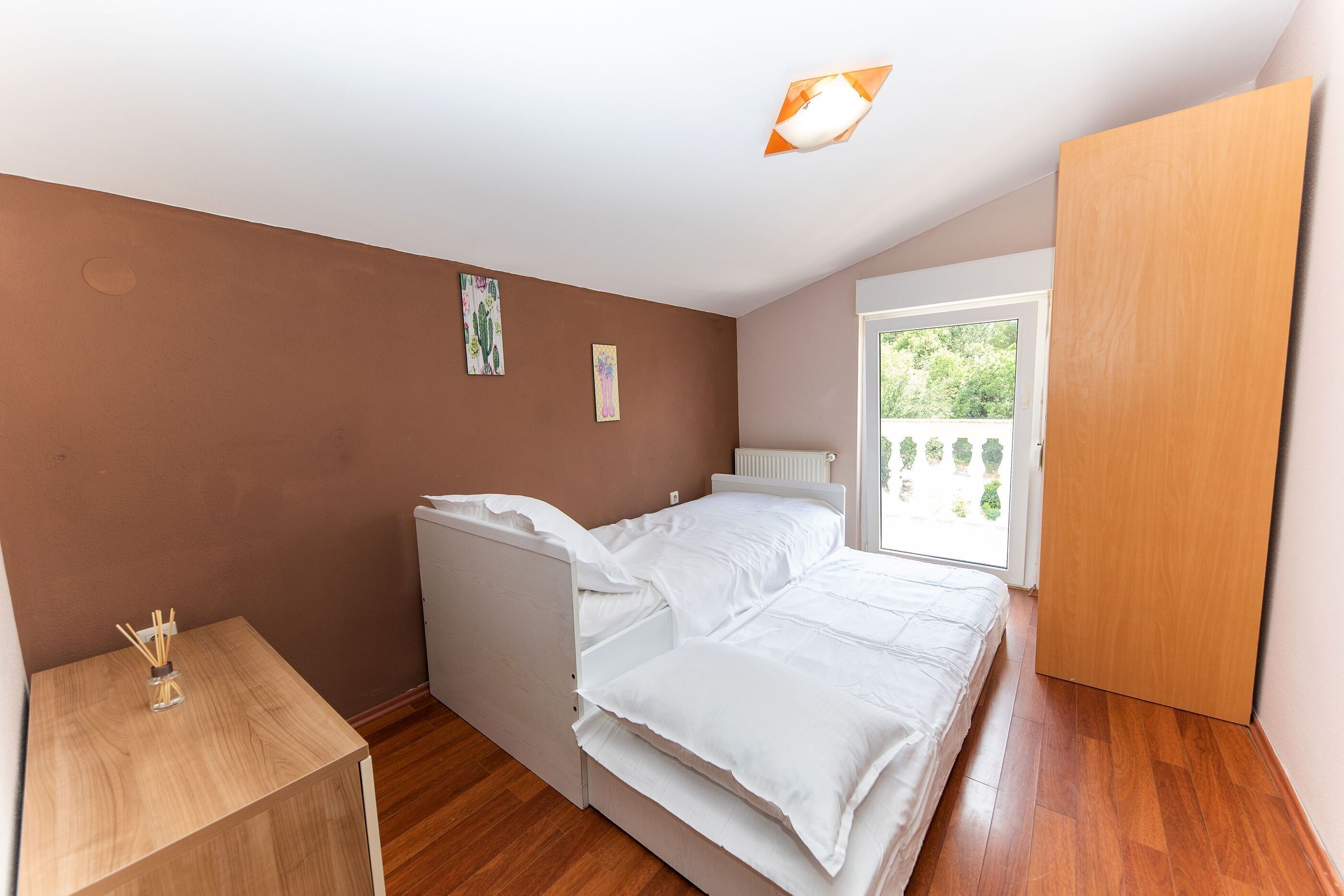 A bedroom with a bed, a small wooden table, and a window with a view of greenery.
