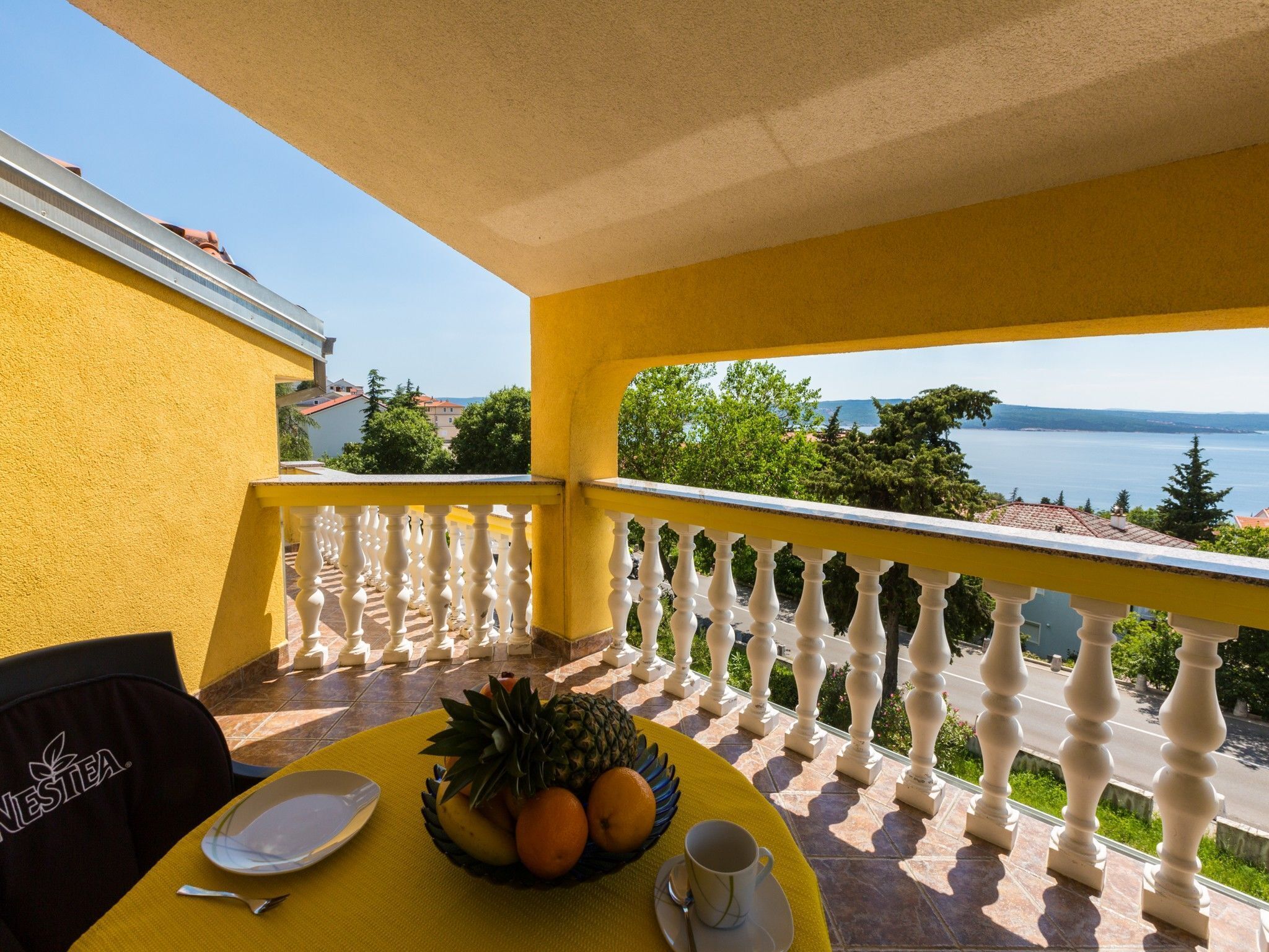 Un balcon jaune avec une table dressée pour deux personnes, un saladier rempli de fruits et une tasse.