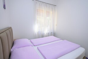 A single bed with a headboard, a window with sheer curtains, and a plain white wall. - Apartments Franco (Selce)
