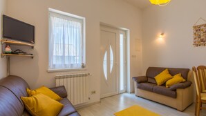A living room with a brown leather sofa, a brown couch with yellow cushions, a yellow rug, a TV on a shelf, and a window with sheer curtains. - Apartments Franco (Selce)