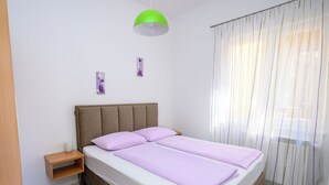 Quarto com cama, mesa de cabeceira, abajur e janela com cortinas.