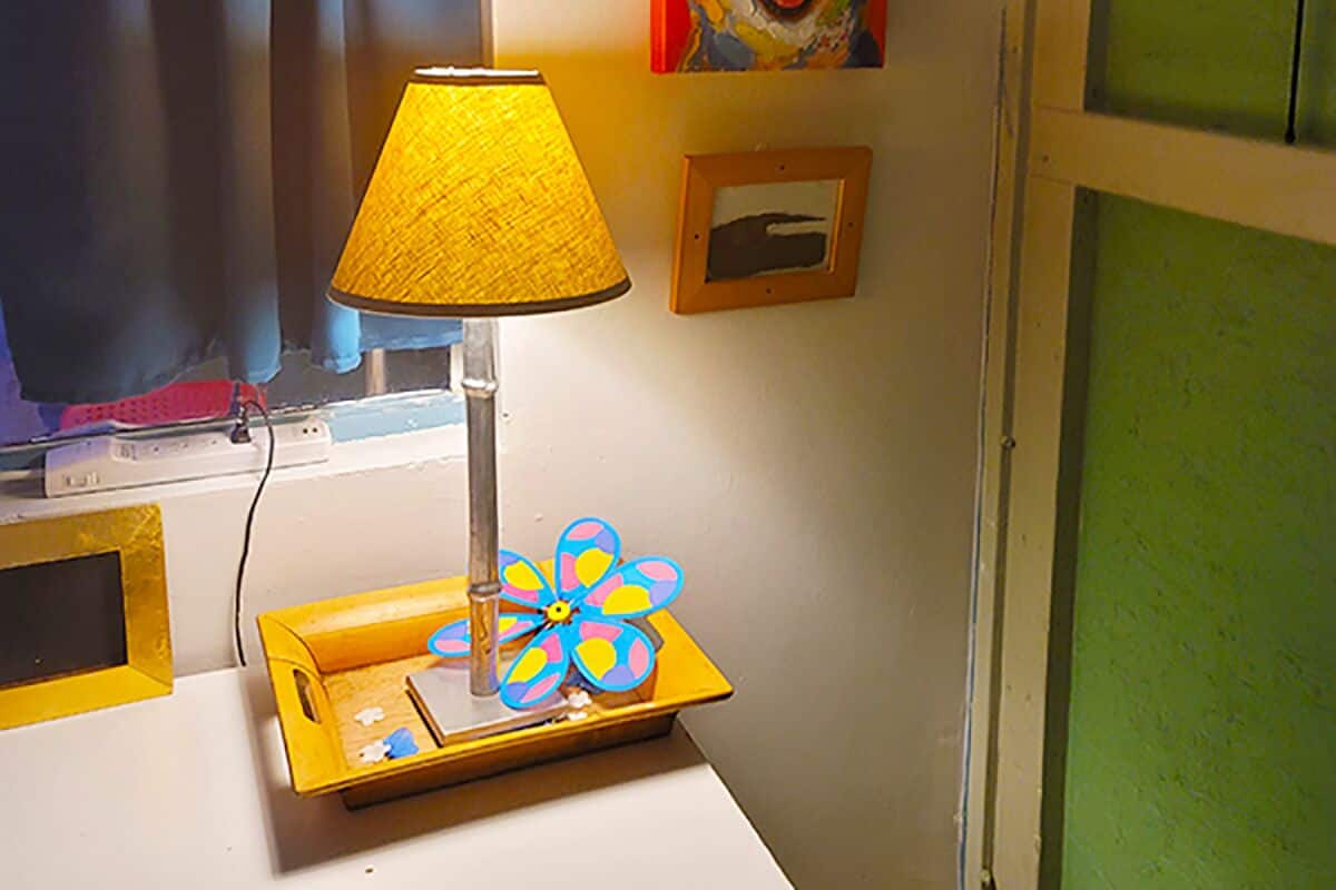 A room with a lamp on a table, a window with blue curtains, and a wall with a framed picture and a pinwheel decoration.