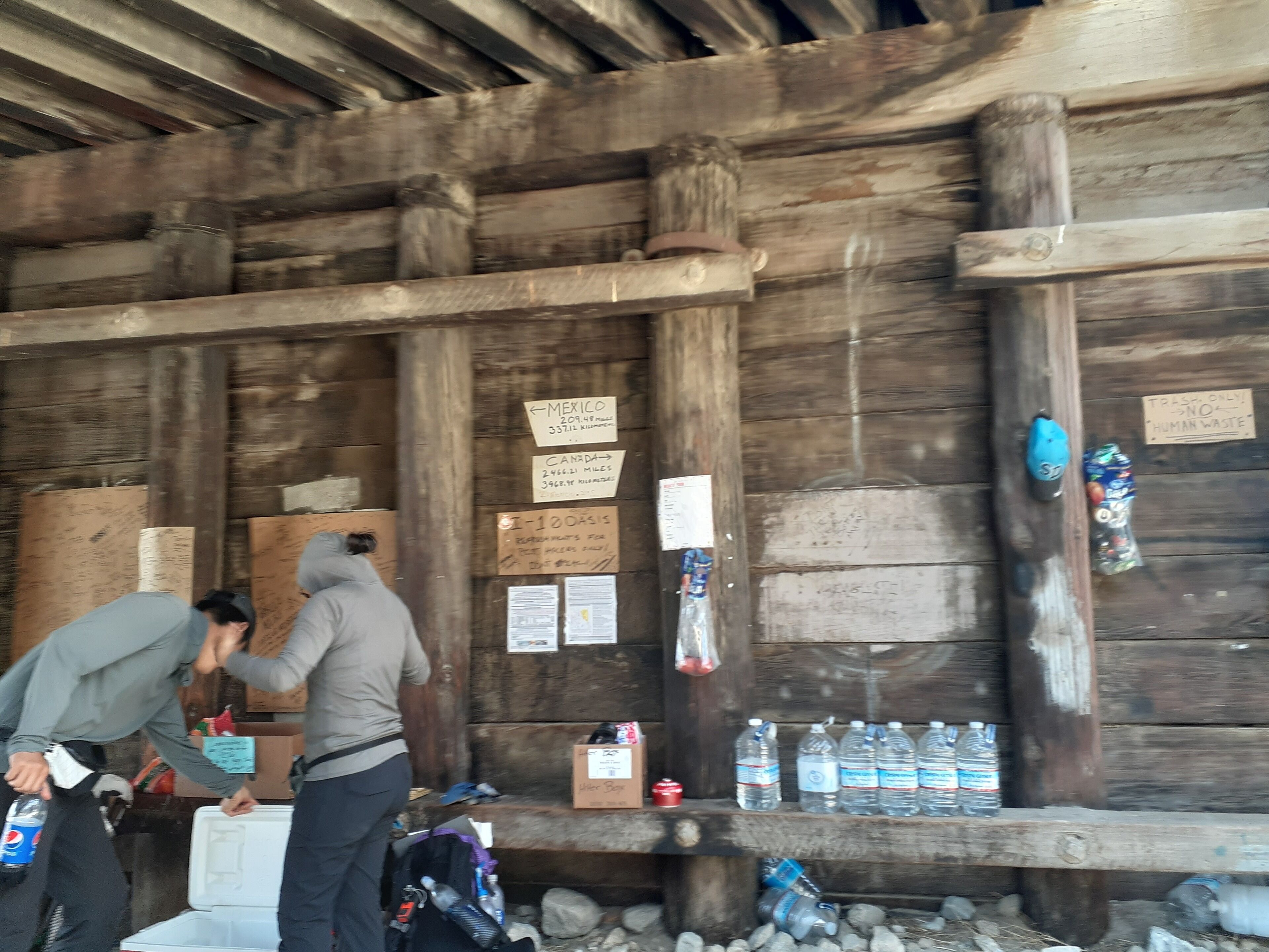 A rustic wooden cabin with a wooden ceiling and walls, a bench with water bottles, and a sign indicating a distance to Mexico.