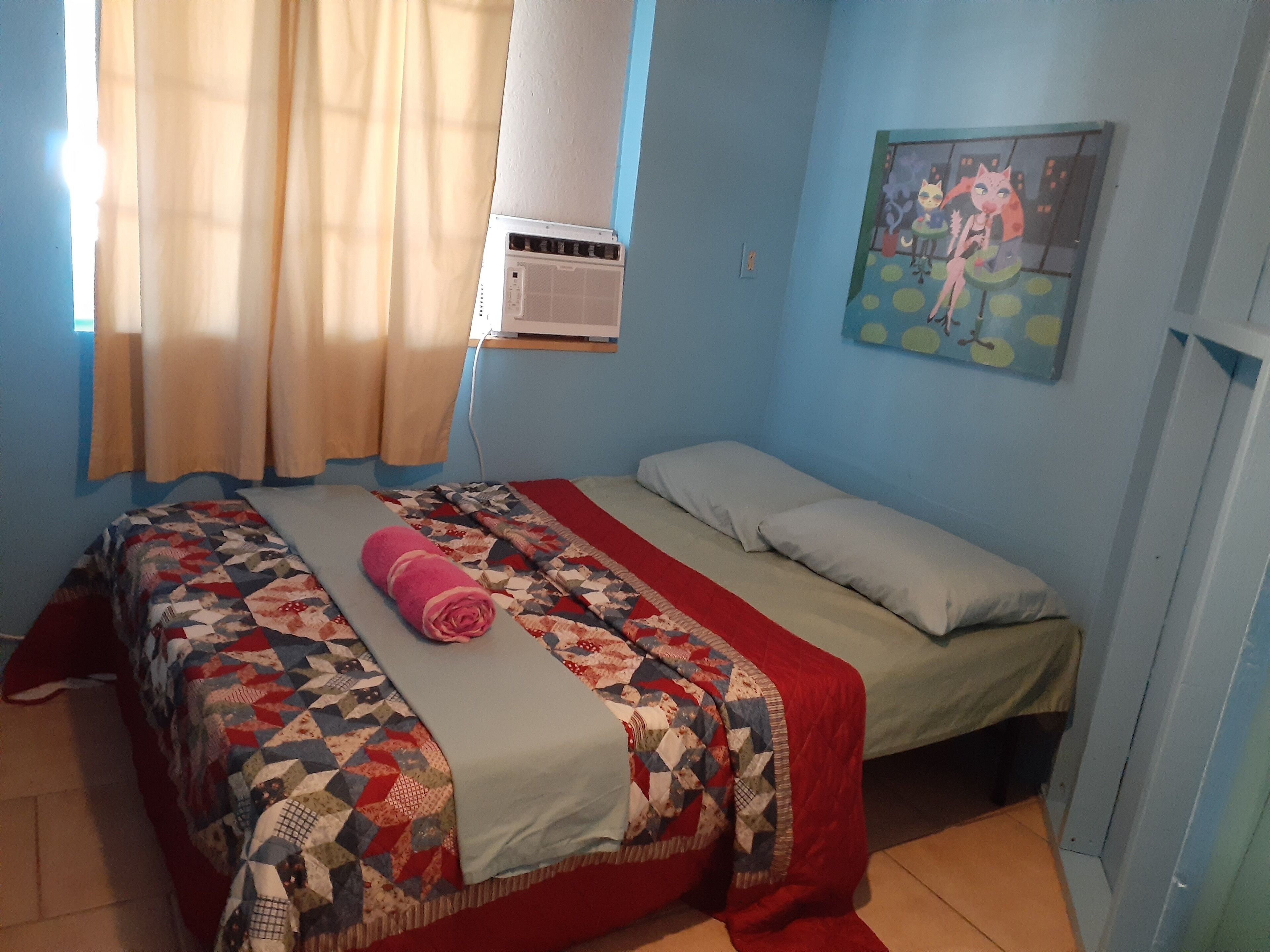 Une chambre à coucher avec un lit, une fenêtre avec des rideaux, un climatiseur et un tableau accroché au mur.