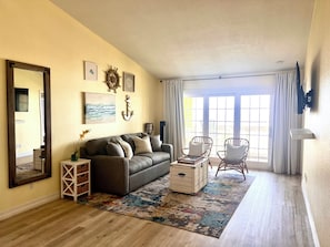 A living room with a sofa, two armchairs, a coffee table, a rug, a side table with a plant, a wall-mounted TV, and a large window with curtains.