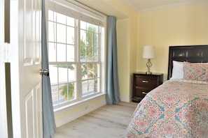 A bedroom with a large window, a bed with patterned bedding, a nightstand with a lamp, and a wooden floor.