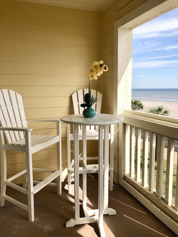 A balcony with two white Adirondack chairs, a small table, and a vase with yellow flowers.