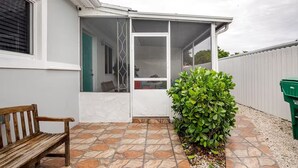 A covered patio area with a bench, a green bush, and a white door leading to a room.