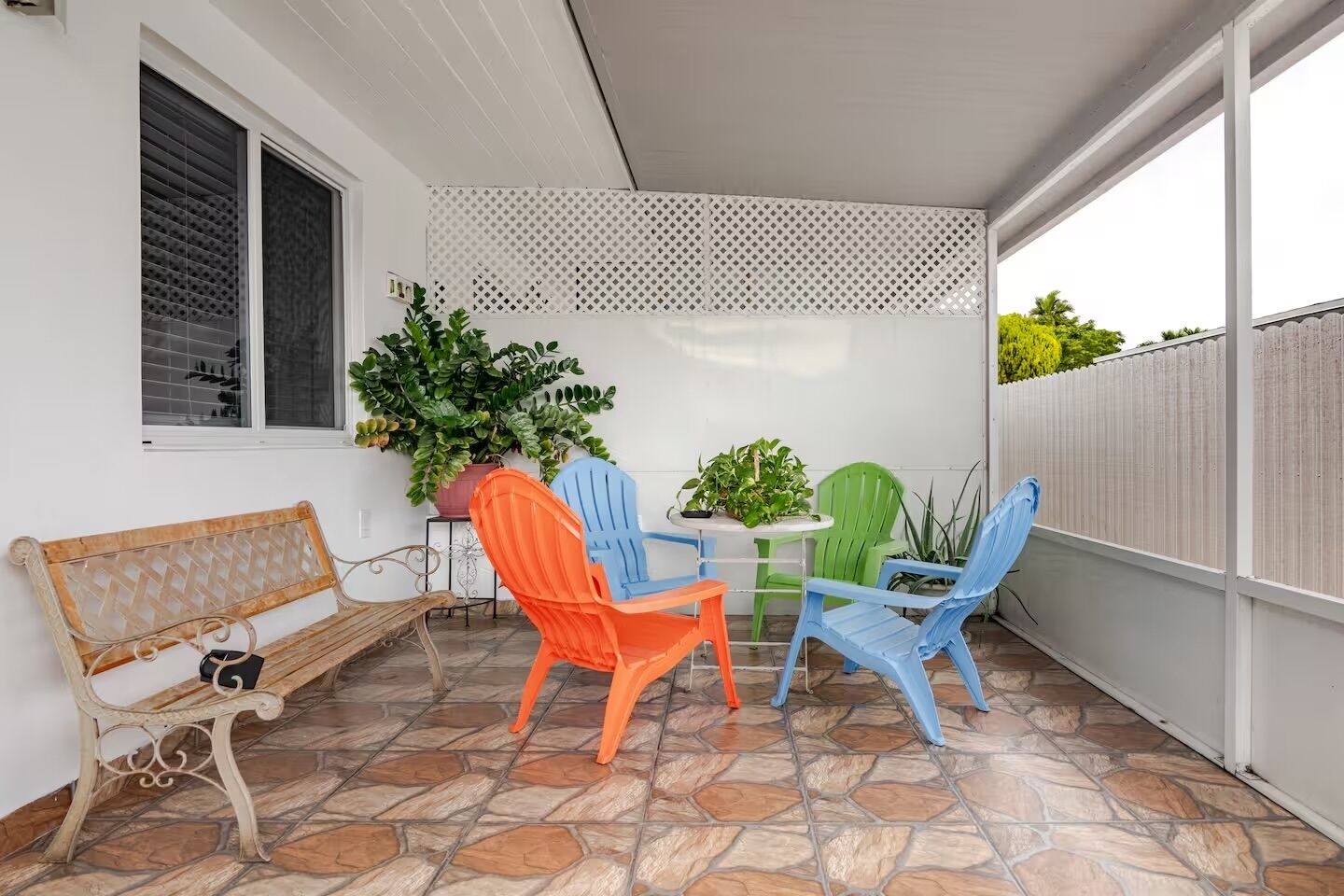 A patio area with a stone-tiled floor, a wooden bench, and colorful plastic chairs.