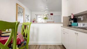 A kitchen with white cabinets, a ceiling fan, and a dining area with a green chair and a table with a plastic bag.