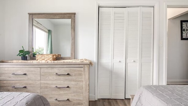 A bedroom with a wooden dresser, a mirror, a closet with white shutters, and two beds with patterned bedding.
