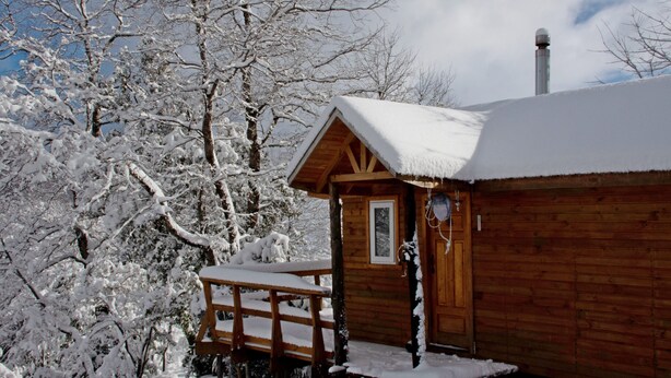 A wooden cabin with a snow-covered roof and a wooden porch. - Conguillio Park Cabin Cabin 3 people (Melipeuco)