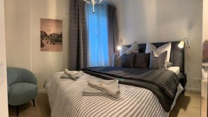 Un lit bien fait, avec une literie à rayures, une lampe de chevet et une chaise, dans une pièce dotée d’une grande fenêtre et d’un tableau au mur.