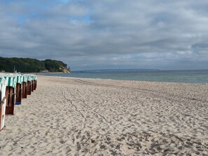 Une plage bordée de rangées de chaises longues et de parasols, un rivage sablonneux et une vue sur la mer et la terre au loin.