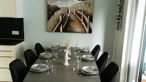 Un coin repas avec une grande table dressée pour six personnes, entourée de chaises noires, d’un tableau représentant un paysage côtier et d’une fenêtre offrant une vue panoramique.