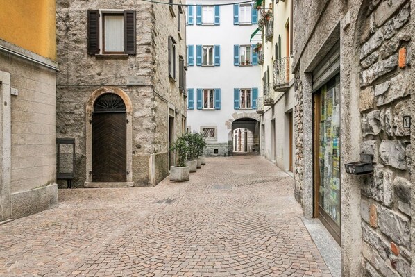 Une ruelle pavée étroite, bordée de bâtiments anciens en pierre, dotés d’arcades et de volets bleus.
