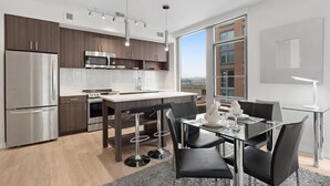 A modern kitchen with stainless steel appliances, a dining area with a glass table, and a large window offering a city view. - Modern 2BR | Full Kitchen | Near Crystal City Shops | By GLS (Arlington)