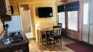 Interior de uma cabana de madeira com cozinha, sala de jantar e televisão na parede.