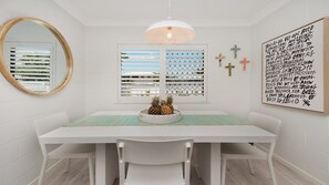 Un coin repas avec une table blanche, des chaises, un luminaire suspendu, une fenêtre avec des stores et un mur orné d’une citation inspirante.