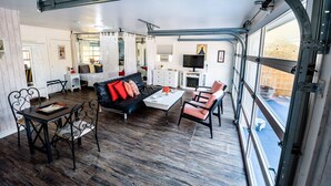 A spacious living area with a black sofa, red cushions, a white coffee table, and metal chairs. A television is mounted on the wall, and there is a large window providing a view of the outside. - Romantic retreat walking distance to wineries  (Temecula)