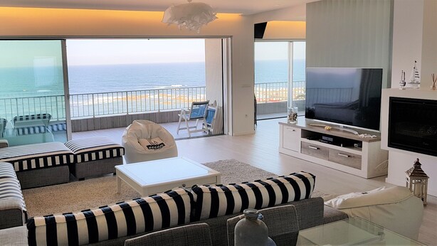 Un salón moderno con televisor grande, asientos cómodos y un balcón con vista al mar.