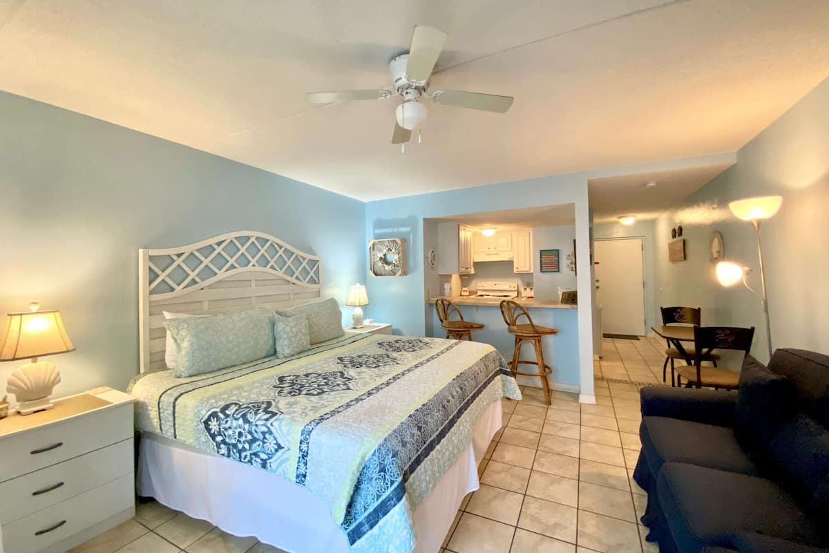 A bedroom with a bed, bedside tables, a ceiling fan, and a kitchenette in the background.