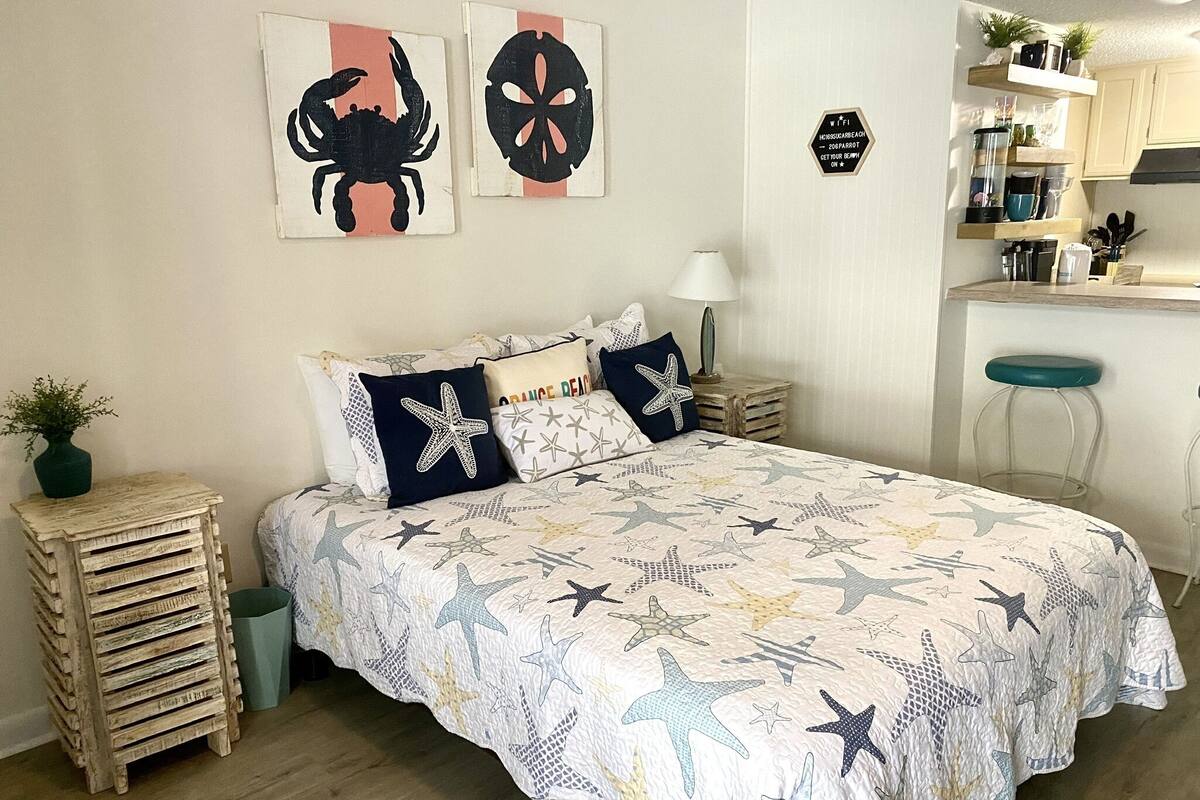 A bedroom with a bed, a nightstand, a small table with a plant, and a wall with artwork.